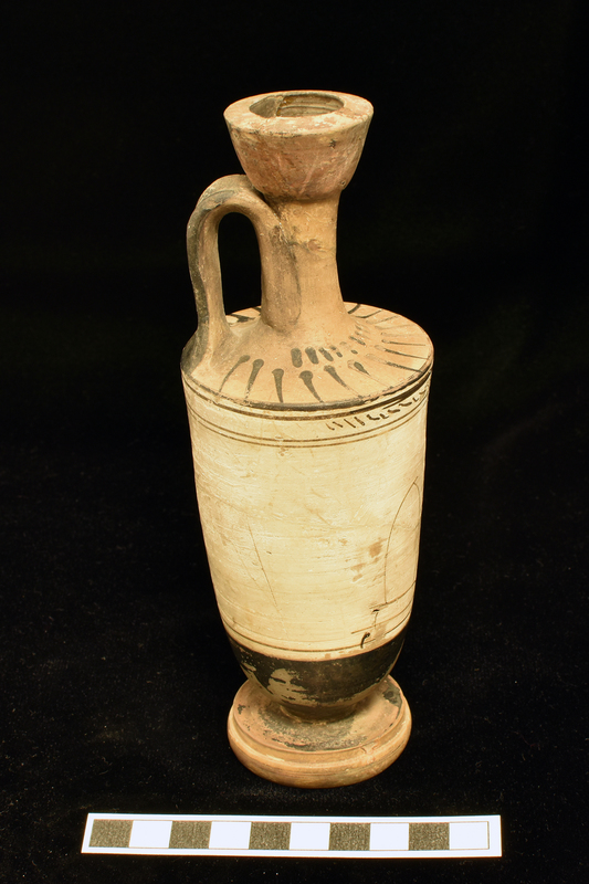 Trumpet-mouth  lekythos with in main white slipped zone opposite handle, badly drawn in dilute glaze, woman in chiton and himation standing before tomb with: 2 stripes below and above 2 stripes and narrow band with irregular diagonal stripes from one side of vase to other above figure d zone.  Lower body, top of base solidly coated, single stripe around top of lower torus of profiled base.  Shoulder decorated with radiating stripes below short band of stripes at base of neck; interior and exterior of mouth and exterior of handle solidly coated.