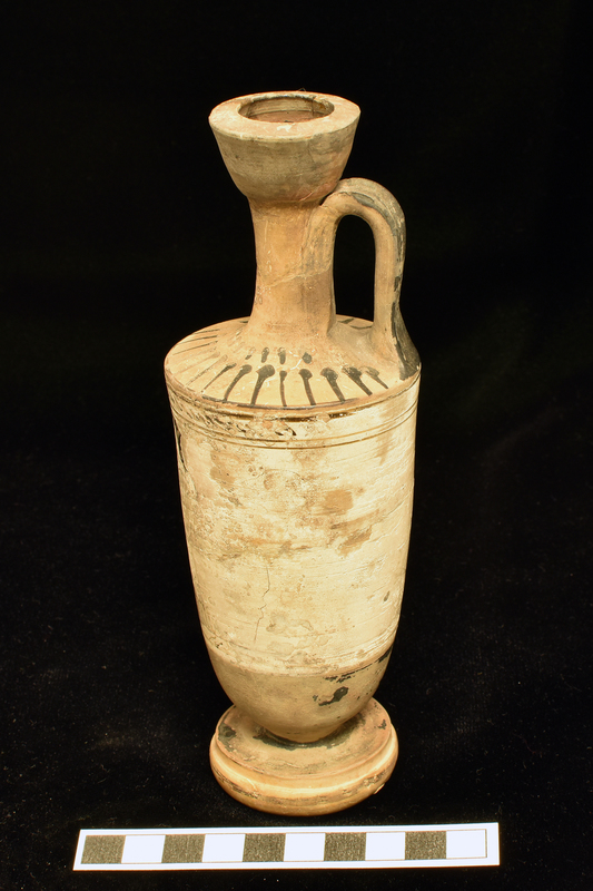 Trumpet-mouth  lekythos with in main white slipped zone opposite handle, badly drawn in dilute glaze, woman in chiton and himation standing before tomb with: 2 stripes below and above 2 stripes and narrow band with irregular diagonal stripes from one side of vase to other above figure d zone.  Lower body, top of base solidly coated, single stripe around top of lower torus of profiled base.  Shoulder decorated with radiating stripes below short band of stripes at base of neck; interior and exterior of mouth and exterior of handle solidly coated.