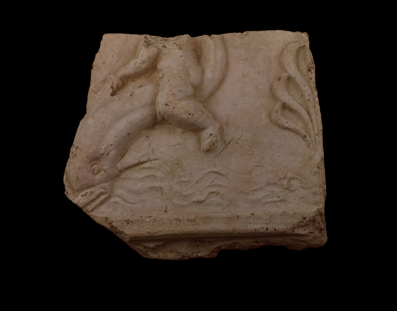 Plaque of medium thickness w relief figure of pudgy child, probably a Cupid, riding dolphin which arches up out of waves; Cupid holds dolphin fin in l hand; features of dolphin’s head well articulated, including ruff of fins behind eyes; light articulation of surfaces of side fins; toes of Cupid articulated.  Waves articulated below dolphin and half of 7-petalled palmette at finished edge behind figures.  Below figures raised ridge of rect section from which projects a bit of finished , squared off lower edge of plaque.  Back flat.
