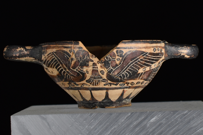 Bowl w convex sides spreading from low offset base to plain rim, prominent nipple on int center; horizontal handles of rounded rectangular section.attached at either side at rim.  Main zone of decoration defined by 2 h stripes above, one below filled w opposing sphinxes on opposite sides of inverted palmette w small blob rosettes in main fields, larger under handles, all decorated w fine incised detail; below rays rise from base which solidly coated on ext w paint continuing onto to underside of base; handles solidly coated on ext.; int thinly coated w paint of varying thickness, so as to produce h stripes.