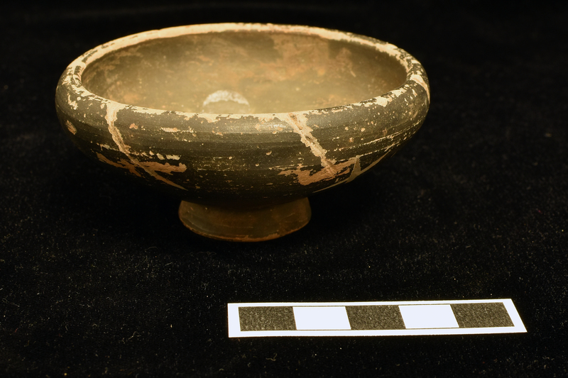 Open bowl with incurved rim on low pedestal base hollow beneath.  Coated inside and out with black glaze.  After firing long diagonal slashes made on exterior from rim; two X’s incised at distance of about 1/3 circumference from one another on exterior at and just below incurving rim.