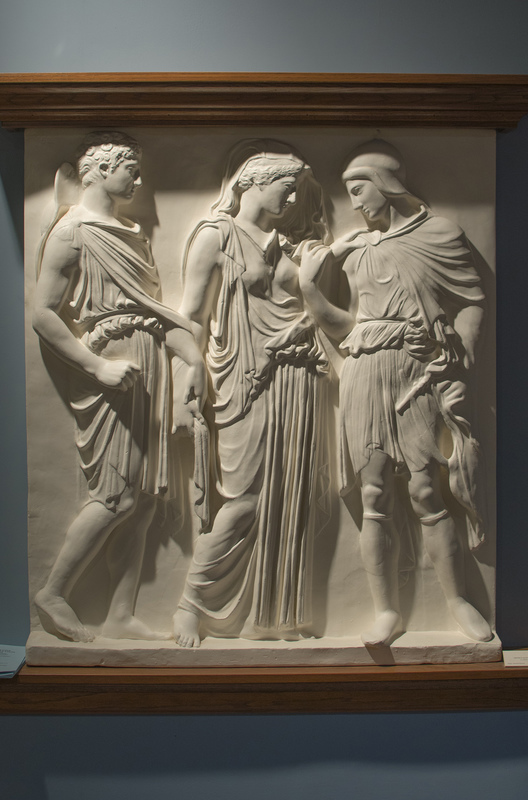 The Orpheus Relief in the Wilcox belongs to a class of so-called "three figure reliefs" in Classical (5th c. B.C. Greek) style. Most of these, however, have been found at sites in Italy, and thus may have been produced by Greek artists for an Italian art market in the 1st c. B.C. or 1st c. A.D. Five full copies and two partial ones are known; our cast is of the relief in the Naples Archaeological Museum, Italy. Inventory no. 6727. Marble. H. 1.18 m. (3 ft.)

The Myth

Our panel shows Orpheus, Eurydice, and Hermes (the Roman Mercury). According to myth, Orpheus was a son of Apollo and a famous singer who lived in Thrace in northern Greece - his song could charm human beings, wild animals, and even inanimate objects like trees and rocks. His wife, Eurydice, was playing with her companions when she stepped on a serpent that bit her fatally. Hermes, conductor of dead souls to the Underworld, led her "shade" into the realm of Hades and Persephone. Orpheus refused to accept the loss, and followed the shade of his wife. Persuaded by his music, Hades agreed to return Eurydice to the land of the living, as long Orpheus, while leading her spirit back to the upper world, did not look back at her. At the threshold to the upper world, Orpheus hesitated and turned - Eurydice was forced to die a second time. In sorrow, the singer turned away from the love of women and became exclusively homosexual. Infuriated, the wild women (maenads) of Thrace tore him to pieces and threw his head in the river; it floated downstream, still singing.

Iconography and Style

Hermes stands on the viewer's left, and can be recognized from his traveler's hat (petasos) and cloak (chlamys) pinned at the right shoulder, leaving the right arm bare. He wears a short tunic belted at the waist. With his left hand he holds the right wrist of Eurydice, who is not only the central figure but also the visual focus of the composition. She stands with her weight on the left leg; the right is free and trailing. Her left hand is raised to the shoulder of her husband, as if to comfort him. Eurydice wears a Classical peplos, a simple dress pinned at each shoulder, with an overfold that covers her belt; over the left leg, the skirt forms closely-spaced vertical folds, like the flutes of a column. A veil completes her costume, and frames her face and downcast eyes. Both Eurydice and Hermes wear sandals with carved soles; the straps would have been painted. Orpheus at right balances the figure of Hermes in a near mirror-image pose (his right hand is raised, however, to touch the hand of Eurydice on his shoulder), and he wears high boots. The singer wears a peaked Thracian cap to indicate his northern origin (his face is restored), and he grasps his lyre in the left hand: the instrument is difficult to recognize, since it appears in profile. The pathos of the scene depends largely on the pose and gestures of the three figures, but as spectators we sense the tragic outcome of the story. L.-A. Touchette, however, has suggested a new interpretation – that Eurydice is shown returning to her husband. The name of each figure is inscribed at the top of the scene in Greek (Orpheus is written backwards, or retrograde). One theory, now discredited, is that the original of this relief once decorated the Altar of the Twelve Gods in the marketplace (Agora) at Athens. More probably, this panel and similar reliefs are much later, and were executed as decorative art for the houses and villas of wealthy Roman patrons. Educated Romans were familiar with Greek myth and could read both Latin and Greek. Perhaps the best-known versions of the story of Eurydice and Orpheus are found in the Georgics of Virgil (70-19 B.C) and in the Metamorphoses of Ovid (43 B.C.-A.D. 17). Orpheus singing is also a favorite subject in Roman mosaics. Much later, he becomes a popular topic in European music and painting.