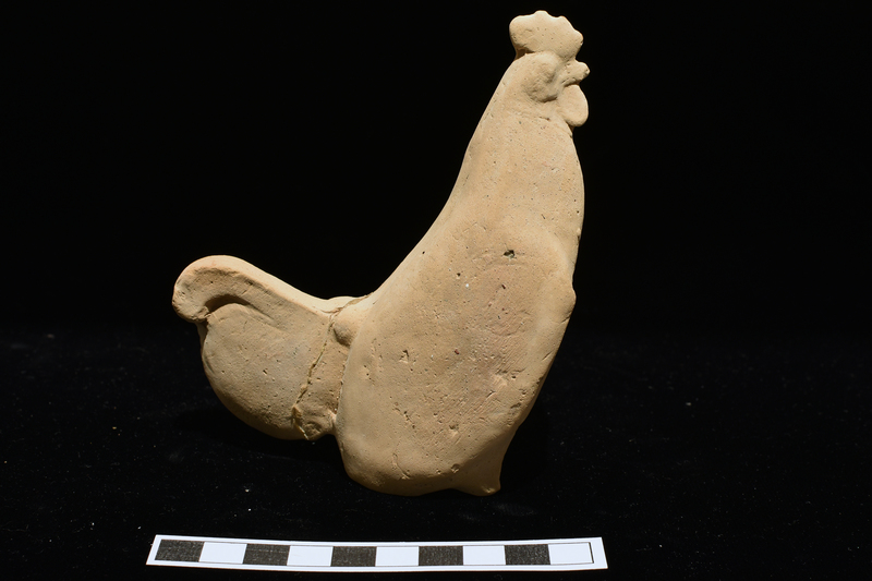 Standing rooster w very short front legs and vestigial haunches providing squarish base.  On head crest, beak, wattles & arms articulated; wings indicated by ridge on either side of body; tail a vertical semicircular projection attached to top of which a curving plume.