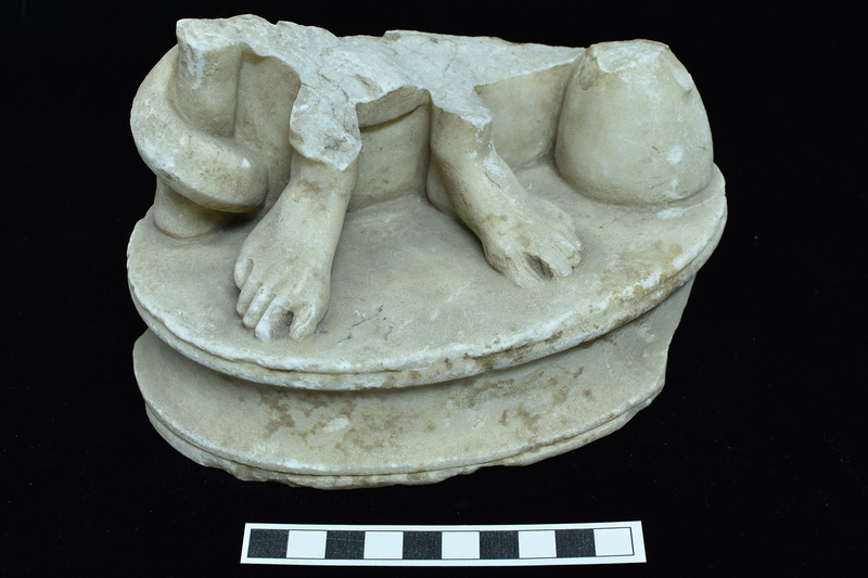 Profiled  oval base with broad central scotia with squared off edge on either side, flanked by torus.  On base two bare feet with traces of garment just above ankles; next to right foot thick gnarled staff with coil of snake which continues behind figure as undulating tail tapering to pointed tip; near left foot cupping vessel.  On feet toenails and joints articulated; horizontal fold articulated on drapery above right ankle.  On underside of base broad roughly smoothed band around edge with center slightly recessed and rough pecked.