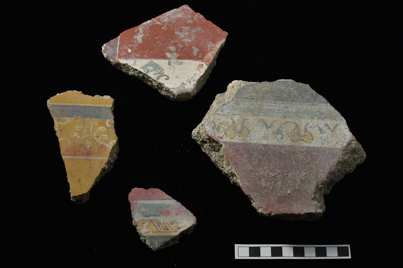 Thick fragment with originally large panels of solid black and solid red on either side of decorative border consisting of band of solid green and slightly broader band painted white with overlaid decoration: abstracted lotus and palmette: lotus flower rising from flanking  leaves of green, floral element outlined with narrow black stripe which curves out to form stem of leaves; palmette reduced to a V-shape done in black paint.  Back surface probably originally flat, now lumpy and irregular in most places.