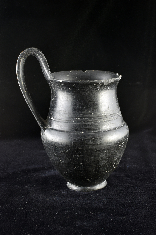 Funnel-necked jug w outturned rim & high-swung flat handle on low offset base; strong incurve of body to juncture w neck; 3 incised horizontal bands on lower part of neck.  Burnished inside & out.