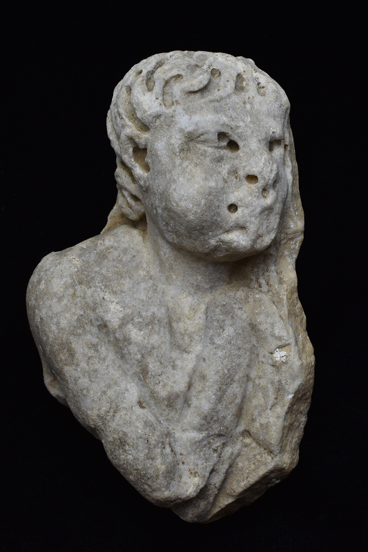 Fragment of upper part of table leg w head & shoulders of male figure w chubby cheeks facing r w upper r arm pulled across body, wearing short-sleeved tunic w roughly V-shaped folds coming from neck; short curly hair articulated with much deep drilling; ears , eyes & mouth articulated.  Back flat.