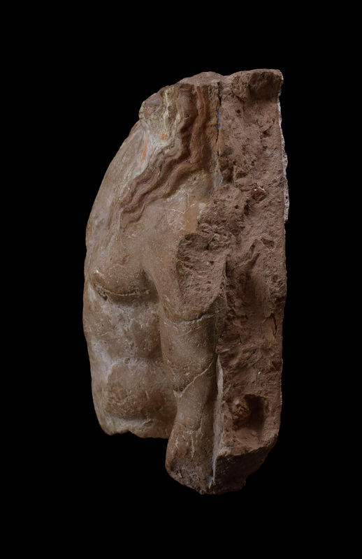 Slab w richly-modeled nude male torso, probably Apollo, w three tendrils of wavy hair falling over each shoulder, r arm held closer to body than l. Coated w white slip w hair painted red and background blue.
