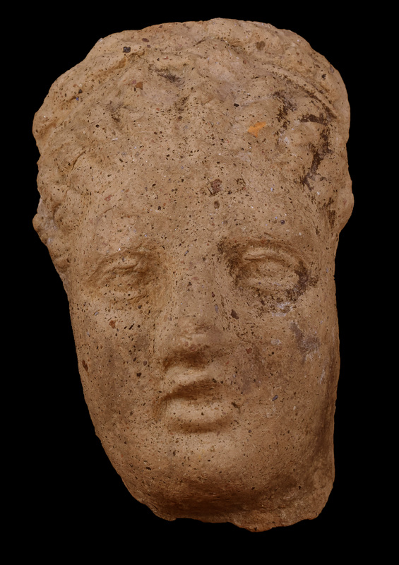 Head with melon hair-do rising from forehead to fillet tied in Hercules-knot with horizontal braid above.  Brows, lids, and pupils articulated; prominent nose above poorly fashioned open lips; fairly broad surfaces of cheeks, prominent rounded chin.  Traces of white slip all over, traces of black paint on hair.
