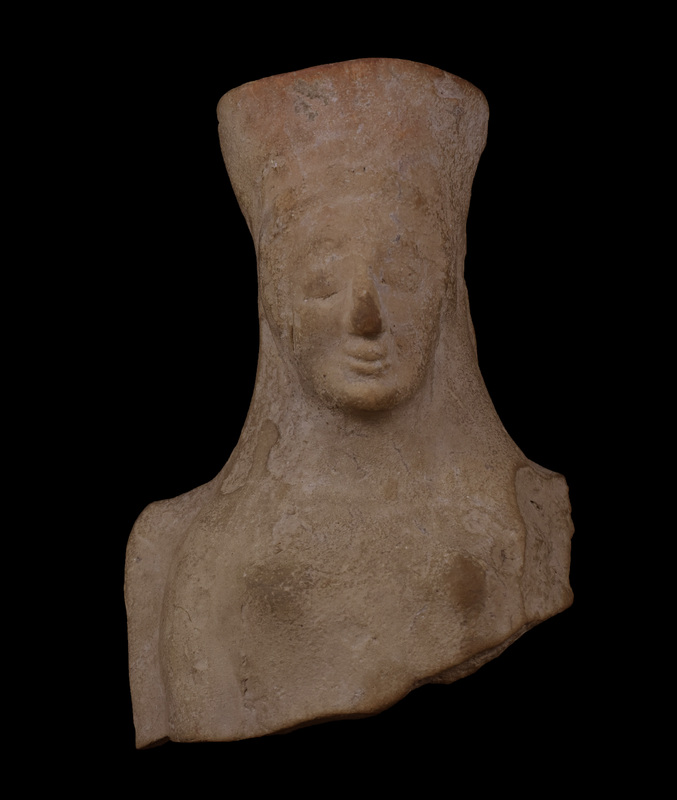 Head  & upper body of woman in polos w slight ridge indicating hair mass above forehead, bulging eyes below ridge of brow, prominent, small upturned mouth w faint “archaic smile.”  Surface of upper body plain with only raised breasts articulated.  Plaque edge squared off behind shoulders.