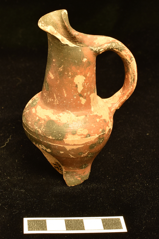 Votive jug with funnel neck ending in broad spout with inturned sides; body with high strongly curved max D from which sides curve inward to slightly depressed underside of base; handle of elliptical section from back of neck to max D.