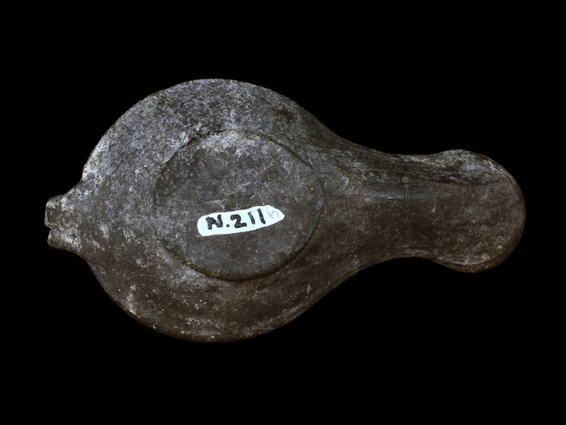 Sharply carinated reservoir w flat discus w fairly small fill hole  surrounded by two raised ridges, bipartite high loop handle fr carination to edge of discus, nozzle w large circular wick hole.  Shoulder decorated w relief  band of ivy leaves & surface of nozzle w ?ax tied w ribbon.  Now all dark gray, ?paint and/or firing.