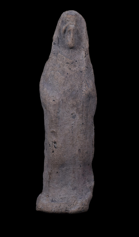Mouldmade standing figure  wearing small polos over which draped palla which falls down along shoulders and is probably pulled  across front of body over slightly bent right arm; apparently palla held at left side at front of body by raised left hand, fingers of which crudely articulated.  Beneath polos hairline articulated and front of face crudely pinched forward; feet rest on shallow base which is bent up from the back and is not level, so that the figure cannot stand unsupported