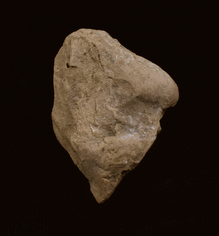 Fragment of figure, possibly male, seated on squarish ?throne.  Only recognizable portion of figure preserved is right arm bent up from elbow, possibly covered by some kind of cloak.