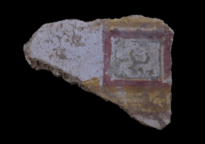 General:  Thin fragment w white background on which painted pair of broad yellow ?v/ stripes, one of which overlaid w brownish triangles,  between which ca square frame composed of concentric stripes: broad red, narrow yellow, possibly originally overlaid w red, broad green with annular green oval in center.  Traces of contiguous square on one side & curving black stripe at one broken edge. Back flat w couple of chunks of wall adhering.