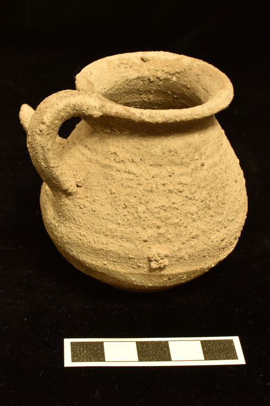 Slightly depressed globular body w slightly concave flat base and outturned rim; long uptilted conical feeding tube attached at just above max D; pairs of horizontal grooves just below rim and on body at level  of spout; vertical handle of rectangular section from just above max D to rim.  Traces of  dripped paint on lower body.