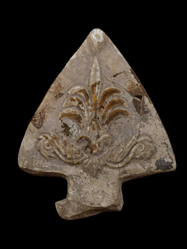 Triangular handle ornament decorated w relief 7-petaled palmette rising from floral base in 2 parts: small lotiform device at base of palmette from which leafy scrolls extend out to near edges.  Originally painted all over exterior with paint extending onto back.