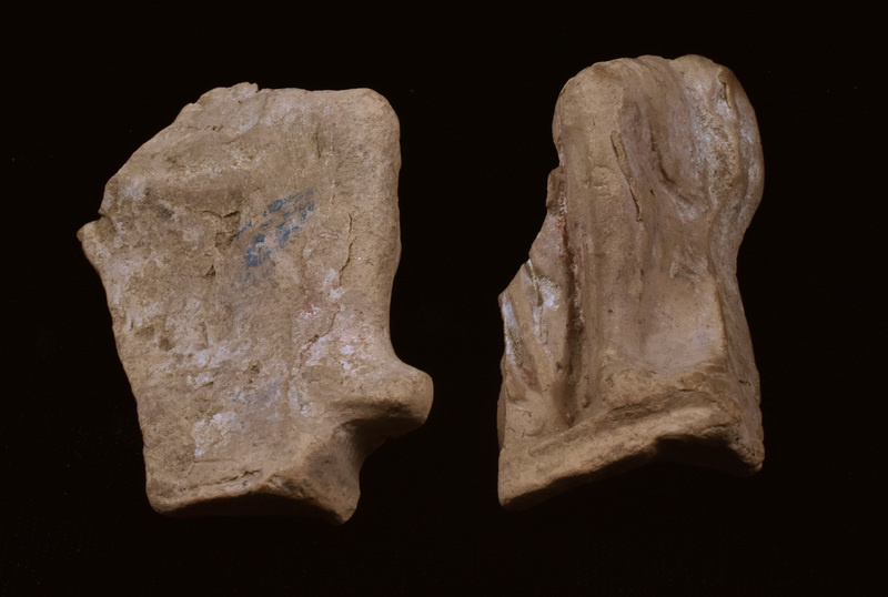 Two fragments of legs of seated figure with drapery folds falling between legs; traces of red and white paint.  Fragment A: bent r leg, possibly w drapery pulled tight over leg; foot projecting in front of squarish base. Fragment B: l leg & base as on A, w traces of red and white paint to r of leg; crude articulation of toes.  On outside of leg traces of drapery over thigh and projection, possibly of seat or throne. Two non-joined fragments which appear to join to inside of right foot; large fragment missing between upper legs, including folds of drapery.