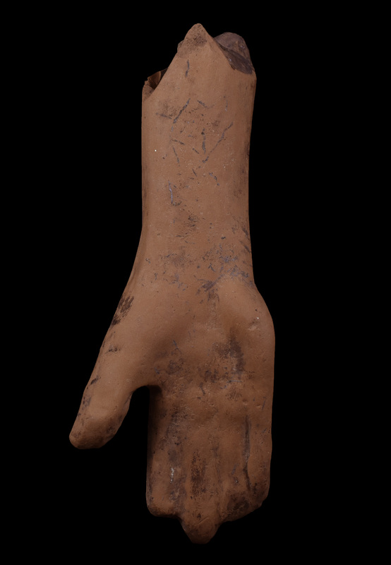 Upraised r hand and forearm.  Crude version of typical form ; no articulations except for thumbnail on lumpy thumb, prominent outer palm pad; wrist of irregularly elliptical section.  Traces of white between fingers probably incrustation & not slip; some dark incrustation.