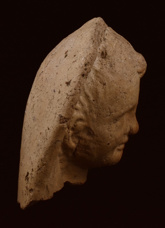 Head of woman in modios, on which discoid pellets, three at top edge, four on either side at bottom edge, between which small pellets with indentations, probably meant to represent pomegranates.  Face of mature woman framed by mass of hair with eyes, nose and mouth clearly articulated;  earrings of disc w  pendent triangular element.  Hollow with top of crown open.