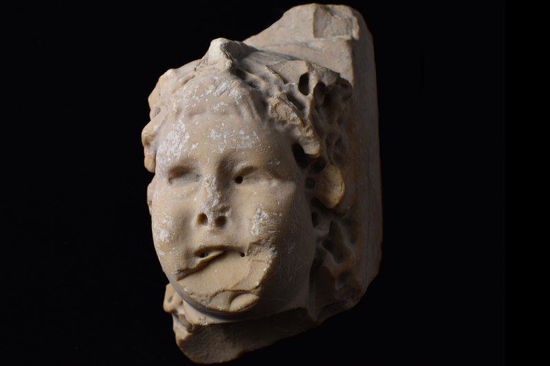 Fragment w frontal face of chubby Cupid w long flowing hair parted in center w central braid from forehead to rear; hair articulated by deep drilling from surface; eyes deeply recessed & deep drill holes at nostrils & ends of mouth.