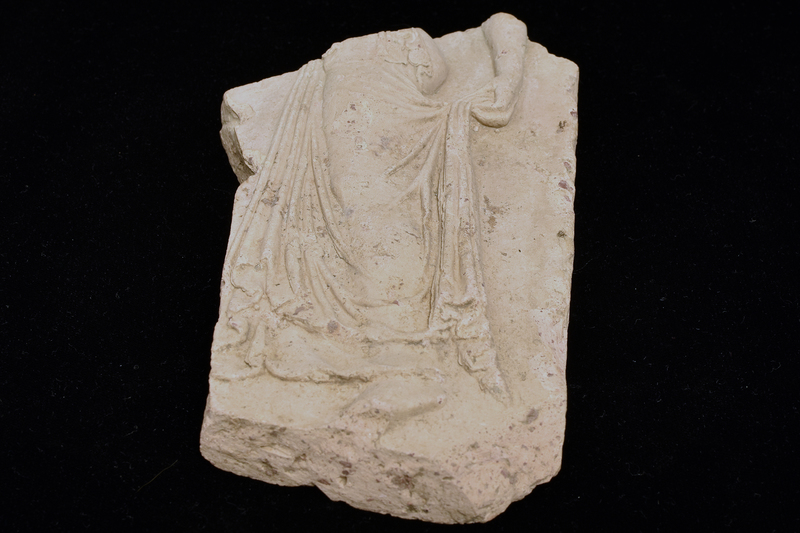 Slab of medium thickness w woman (?maenad) in relief facing l, standing on tiptoe & wearing peplos w long overfold reaching to hip; lower skirt of peplos  pulled across leg to clump of folds held by left hand below buttock & falling along back of leg; fuller mass of folds in front of body, presumably held by missing r hand; folds articulated in archaistic version of late 5th c “wet drapery” tradition.  Back flat.