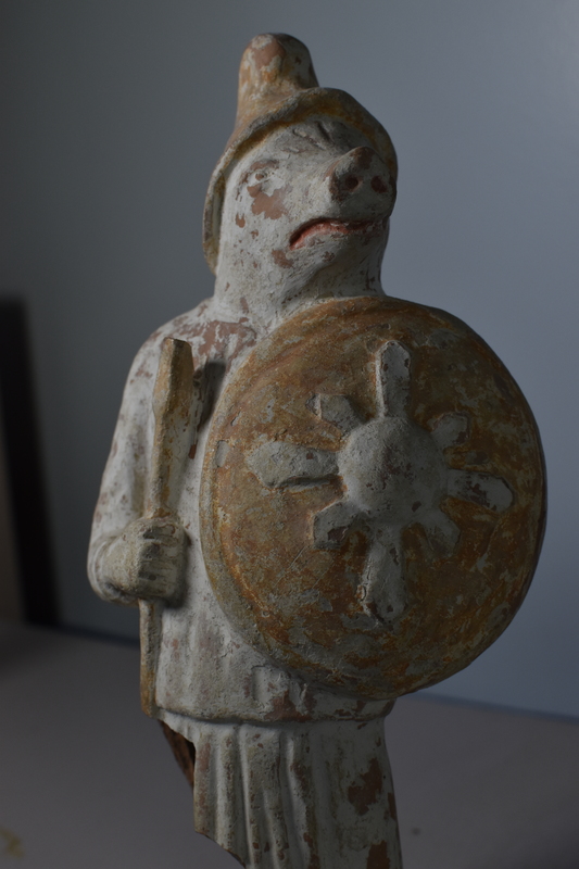 Mold-made figure of standing pig wearing long-sleeved tunic with overfold below waist level & soft-peaked Trojan cap and holding spear in right hand and shield in left, both held close to body; on head, turned to left, eyes, vertical wrinkles on “forehead” and horizontal wrinkles on snout articulated.  On shield, central boss from which extend 8 rays, alternating larger on major axes & smaller between, indentation marking shield rim.  Vent hole at back.  Whole covered with white slip; body of shield, spear, and cap yellowish brown, nostrils and lips red.