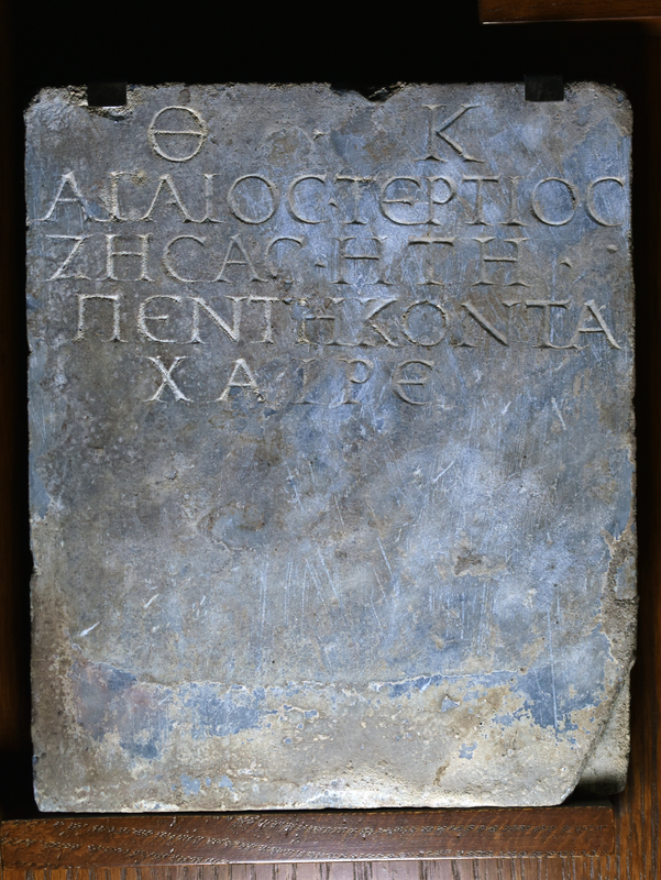 Rectangular slab w inscription in capital letters in 5 lines:

Θ • Κ 
ΑΓΑΙΟΣ ΤΕΡΤΙΟΣ
ΖΗΣΑΣ ΗΤΗ
ΠΕΝΤΕΚΟΝΤΑ
ΧΑΙΡΕ

To the gods of the underworld (θεοῖς καταχθονίοις / theois katakhthoniois)
Agaios Tertios
having lived 50 years
Greetings!

Only face well smoothed, back rough pecked, edges incompletely ground.