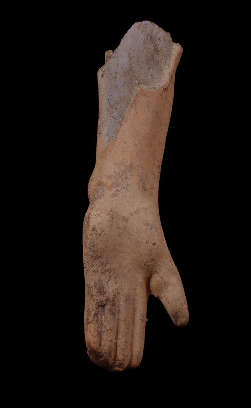 Upraised left hand & forearm.  Four contiguous, unarticulated straight fingers w thumb projecting diagonally to side, thick thumb pad & prominent interior wrist bone; back plain and unmodulated; flaring arm of plano-convex section flattened on sides.  Fait traces of red paint over white slip.