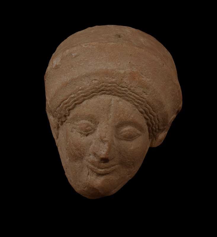 Female in polos w hair above forehead in two masses articulated  w close-set waves.  Slightly bulging eyes w rims articulated above & below under slightly raised edge of brow.  Prominent nose which squeezed while clay still pliable w fingerprint on l side; upturned lips in faint “archaic smile.”  Poorly articulated ears w trace of large pendant earrings.  Suspension hole in top of polos.