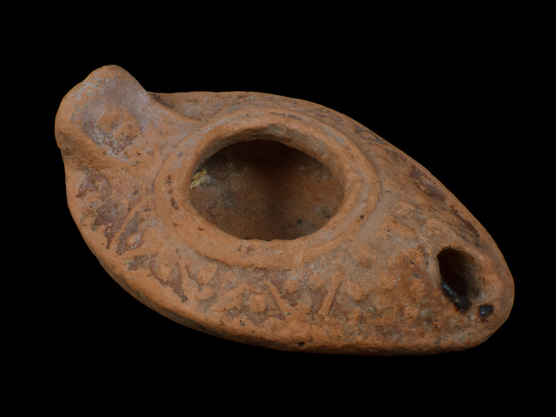 Slipper lamp w upturned solid rectangular lug handle, edges of which defined by relief band within which relief X below, relief ?rosette above.  Carinated reservoir w flattened carination, large fill hole surrounded by two relief bands;  on upper surface decoration of open V's pointing toward carination within and between which relief dots; relief Greek cross on nozzle.  Ca flat base depressed within poorly articulated raised band; possible mark at edge of band two indistinct depressions.  Top surface coated w red paint, some of which dripped to underside; blackened nozzle.