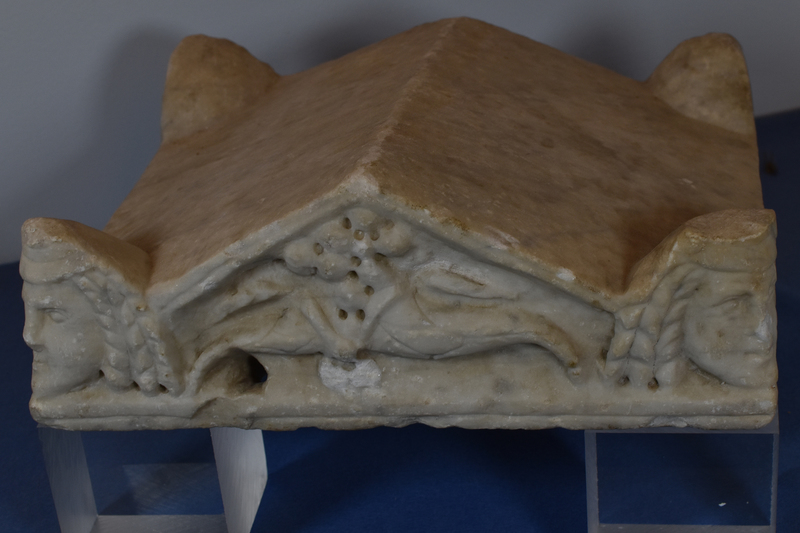 Lid in the form of gabled roof with 4 projecting acroteria at corners.  On primary façade gable filled with two cornucopiae in relief and acroteria carved as dramatic masks: heads, ? male, with crown-like headdress from which 3 long curls extend to frame face on either side; articulation of eyes, nose and mouth.  Gabled surface opposite and acroteria uncarved as are sides and surfaces of gable.