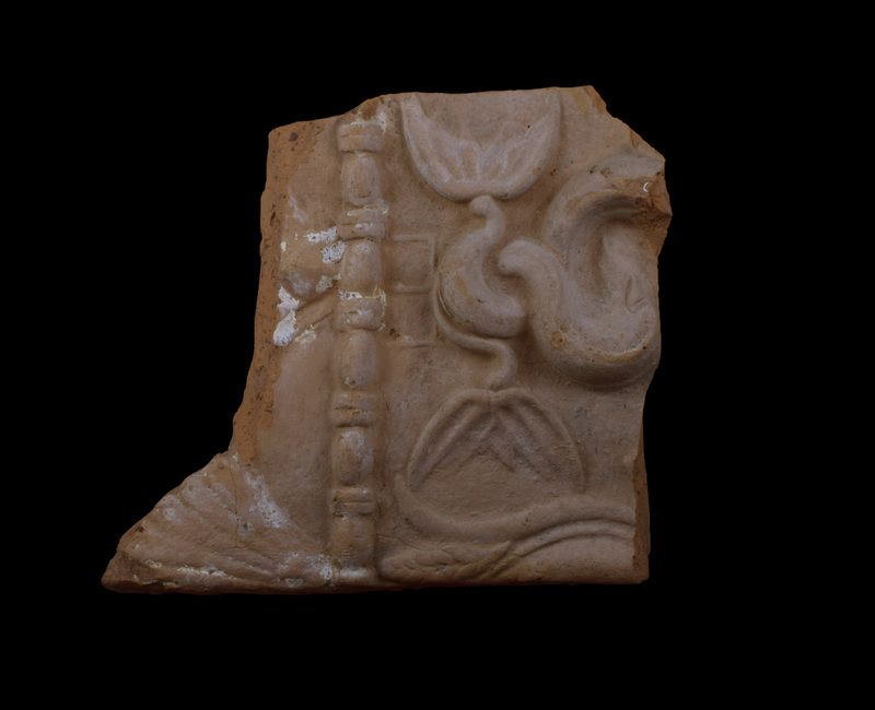 Fragment from top of plaque with h relief band of bead and reel separating top edge from main section of plaque; plaque finished with alternating 9-petaled palmettes and lotus; on main section intertwined tails of fish with opposing tail-fins above which pres base of trident in low relief; at finished edge of plaque part of lotus with a stem of grain between outer petal and central section.  Traces of white slip on front; small patches of white incrustation on front, considerable white incrustation on flat back surfaces.