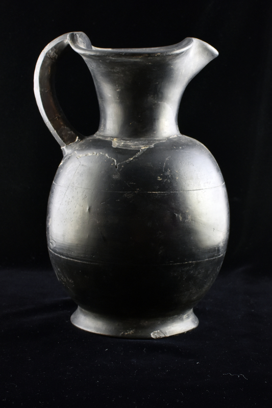 Oenochoe on low pedestal base with handle of pentagonal section from top of rim to top of shoulder; extending from top of handle onto rim horn-like projections from metal prototype; burnished on exterior and interior mouth.