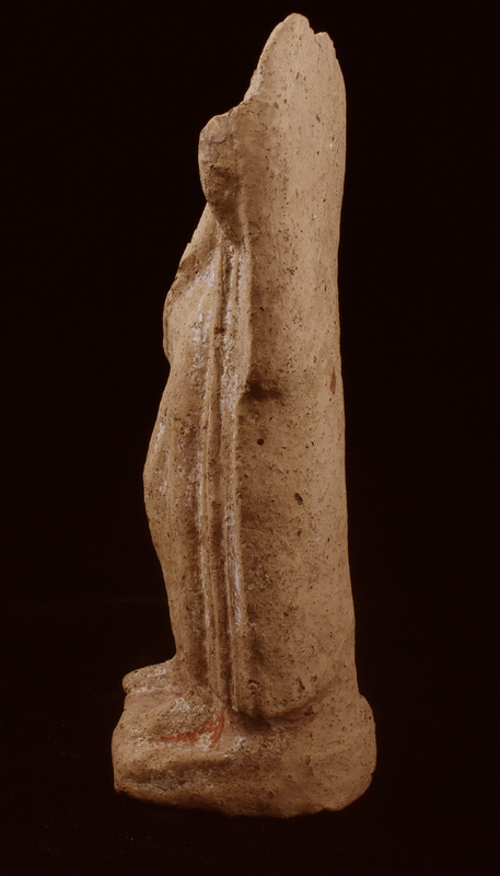 Mouldmade standing figure wearing palla & stola, the former coming around r shoulder and along r arm, pulled diagonally across upper body by raised l arm and falling in vertical folds along left side of body; r leg forward & bent at knee; resting on low base squared at front, rounded at back.  Articulation of top of chiton and plain necklace; chiton folds  vertical in upper half, pulled diagonally by bent leg.  Flat object of uncertain nature rising from l shoulder.  White slip on front & sides, traces of red paint pres only on base.