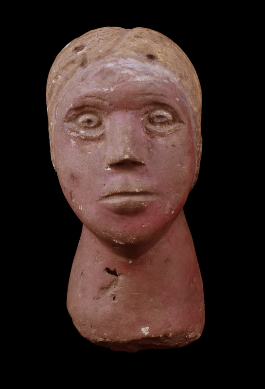 Head and neck of female with wig-like hair parted in middle and pulled back into a bun, hair strands marked by broad sawtooth impressions.  Flat oval face projecting forward with plastic eyes with pupils incised, prominent nose with nostrils indicated, and with horizontal mouth slit on raised surface indicating lips, upper lip with slight horizontal depression at center.  Thick neck spreading toward break, possibly not attached to a body but some kind of base.  Flesh painted reddish, hair showing traces of white paint which is better preserved on forehead in front of hair mass.