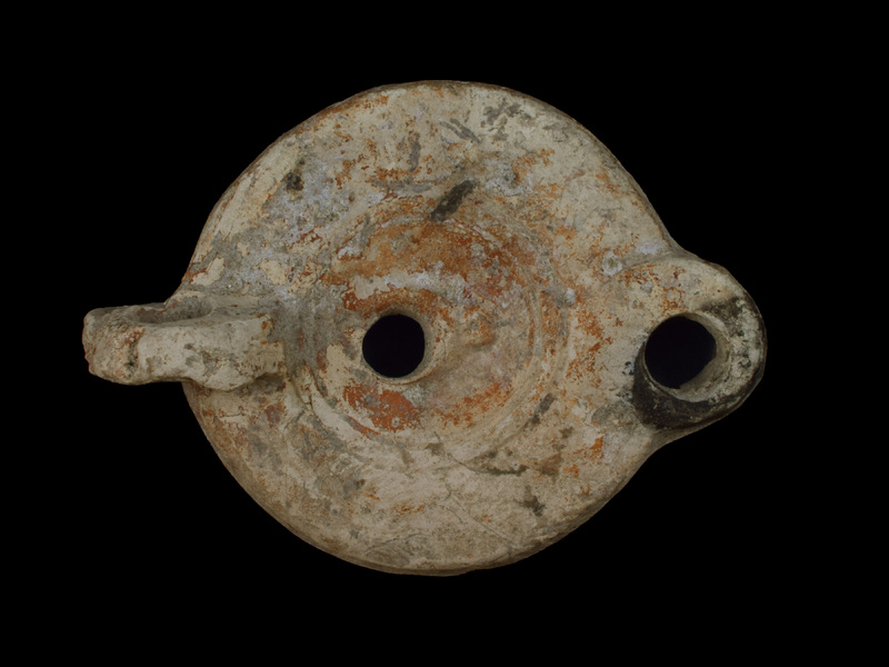 Low convex-sided reservoir w broad, ?plain rim separated from small convex discus by two concentric incised lines; off-center fill hole surrounded by relief crescent moon on ring-shaped base; short rounded nozzle at base on either side incised line & small  incised annulus.  High, horizontally pierced vertical handle from base edge to edge of discus.  Flat base defined by single incised line within which off-center short raised line, tapered at both ends.  Some irregularities, such as depressions in handle and on base, which appear to be pre-firing.  Coated all over w reddish paint; darkening on nozzle.