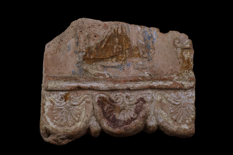 Fragment from bottom of plaque with terminal band of pendant palmettes separated by raised band with central depression from main portion of plaque.  6-leaf palmettes suspended from spiral scrolls which curve up in center, down at ends.  Above raised band heavily draped figure in archaizing style drapery moves on tip-toe toward what appears to be an altar w gabled top & ?lion feet; nature of dress uncertain with swallow tail folds in front of & towards center of figure and at rear stacked folds.  Apparently whole plaque covered with white slip with added red on lower portion of one palmette preserved  and added blue on side panels yellowish-brown.  Yellowish brown on lower portion of altar.  Traces of peg hole on upper edge.