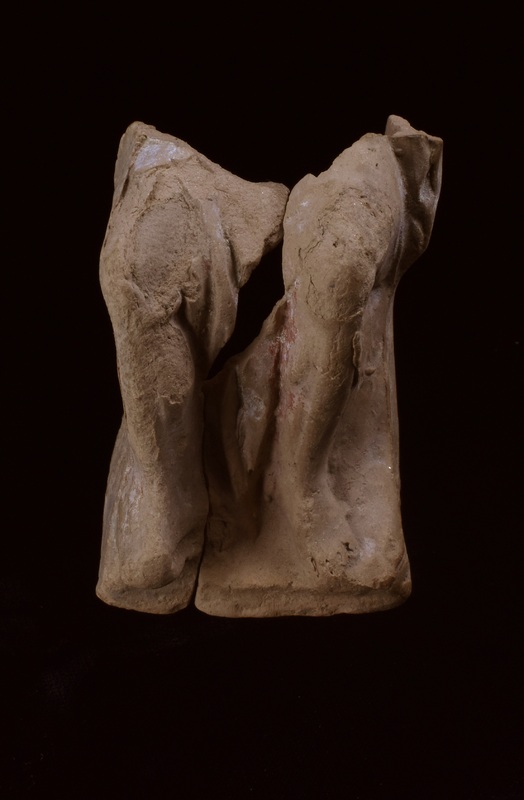 Two fragments of legs of seated figure with drapery folds falling between legs; traces of red and white paint.  Fragment A: bent r leg, possibly w drapery pulled tight over leg; foot projecting in front of squarish base. Fragment B: l leg & base as on A, w traces of red and white paint to r of leg; crude articulation of toes.  On outside of leg traces of drapery over thigh and projection, possibly of seat or throne. Two non-joined fragments which appear to join to inside of right foot; large fragment missing between upper legs, including folds of drapery.