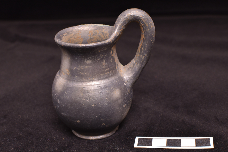 Small jug with offset funnel neck and out-turned rim on low offset base; high-swung handle of circular section from shoulder to top of rim; burnished in and out.