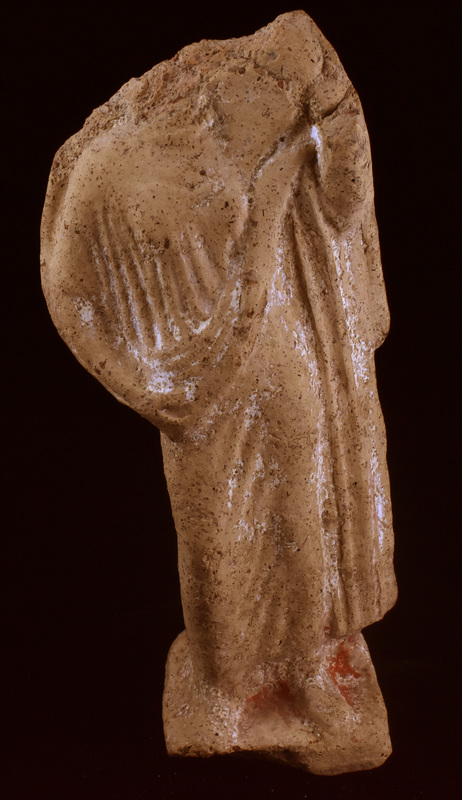 Mouldmade standing figure wearing palla & stola, the former coming around r shoulder and along r arm, pulled diagonally across upper body by raised l arm and falling in vertical folds along left side of body; r leg forward & bent at knee; resting on low base squared at front, rounded at back.  Articulation of top of chiton and plain necklace; chiton folds  vertical in upper half, pulled diagonally by bent leg.  Flat object of uncertain nature rising from l shoulder.  White slip on front & sides, traces of red paint pres only on base.