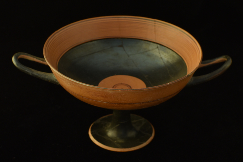 Bowl w slightly uptilted handles on high pedestal foot. Pedestal solidly coated, lower half of  ext bowl decorated w two broad stripes divided by narrow reserved band; upper half divided by narrow painted stripe; handles coated on ext.  Int rim decorated w narrow concentric stripes, bowl solidly coated except for reserved circle in center w narrow concentric stripes in center.