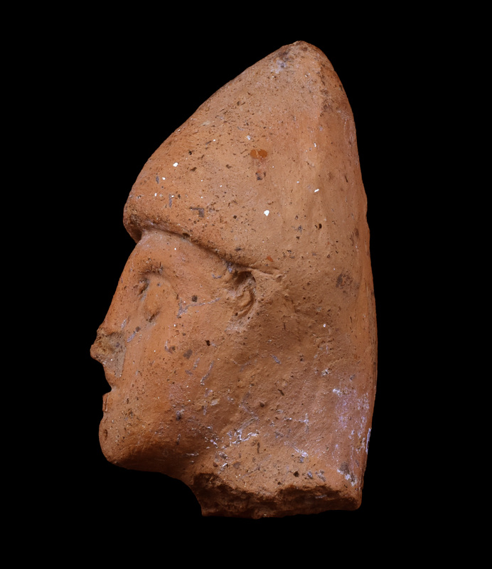 Head of male in pointed cap.  Face triangular with round blunted chin, pellet eyes under overhanging brows, nose modeled, mouth indicated by horizontal incision, ears indicated by small scallop-shape attachments.