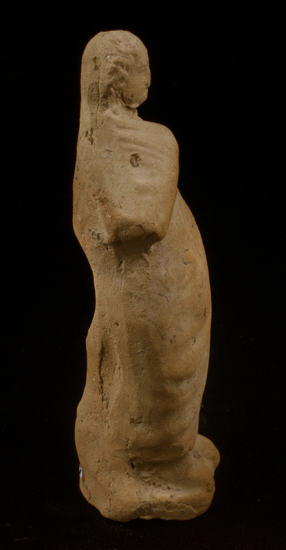 Mouldmade standing figure of Sophoclean type with right arm and hand completely covered by himation and left arm, slightly bent, close to side with large clump of drapery falling forward from left hand; left foot projecting slightly before right, small base.  Eyes, mouth and nose articulated and irregular incisions articulating hair mass which is particularly defined around face.  Very large vent hole in plain back.